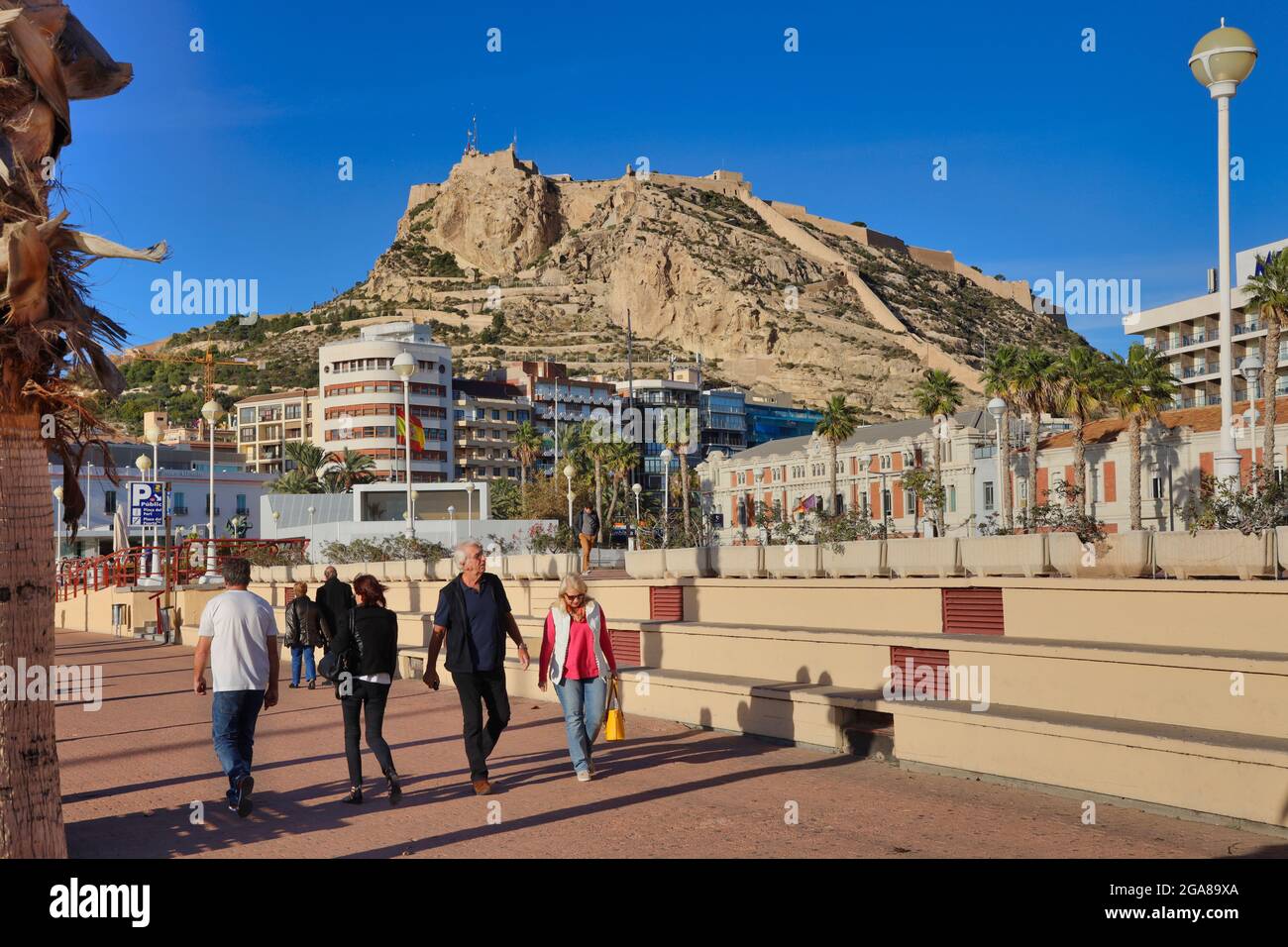 Die Burg von Santa Barbara hoch oben auf einem Vorgebirge mit Blick auf die Stadt Alicante, Spanien, mit der Promenade und den Menschen, die im Vordergrund gehen Stockfoto