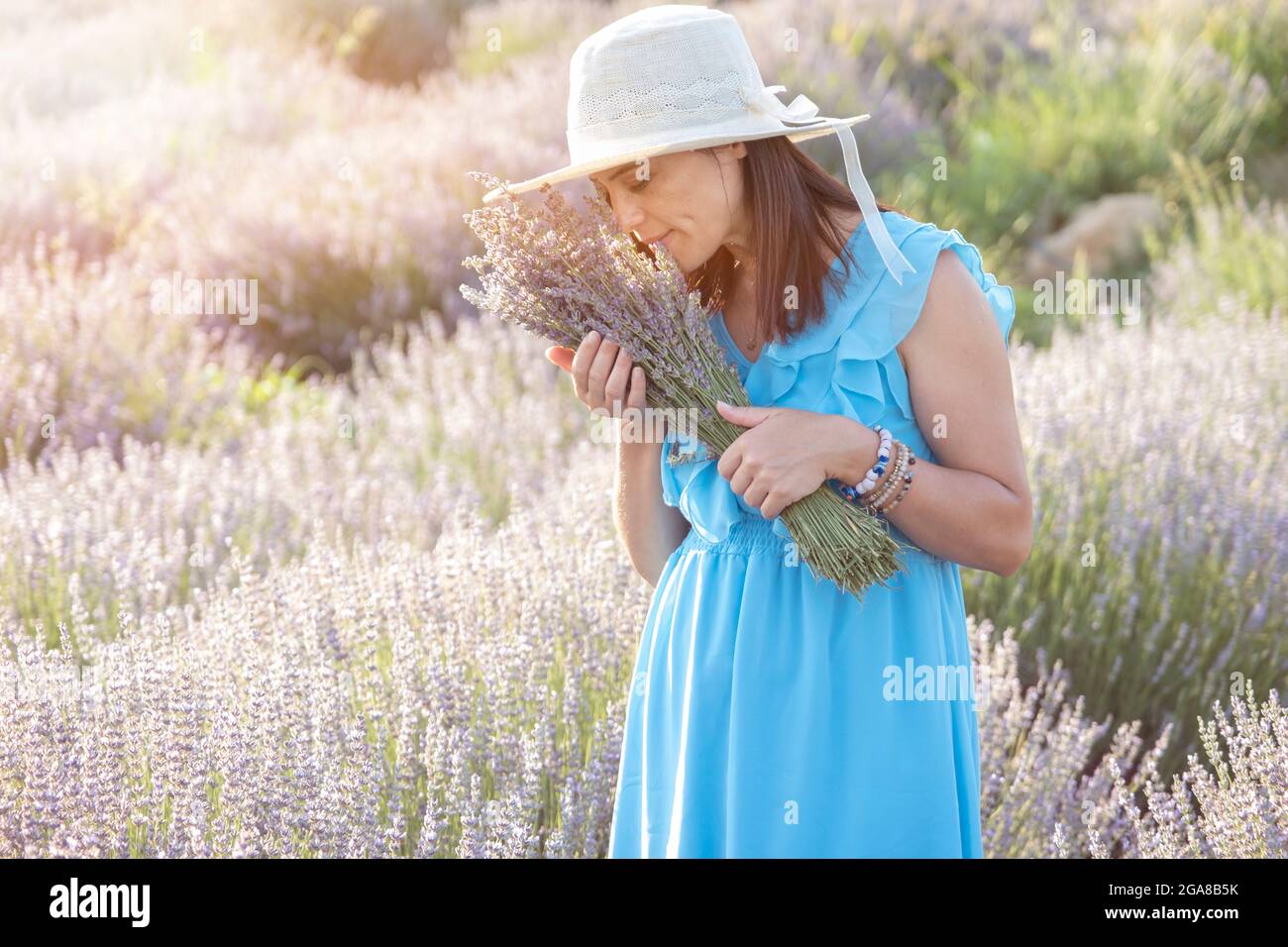 Schöne Schwangere junge Frau in Weidenhut und weißem Kleid posiert in einem Lavendelfeld. Stockfoto