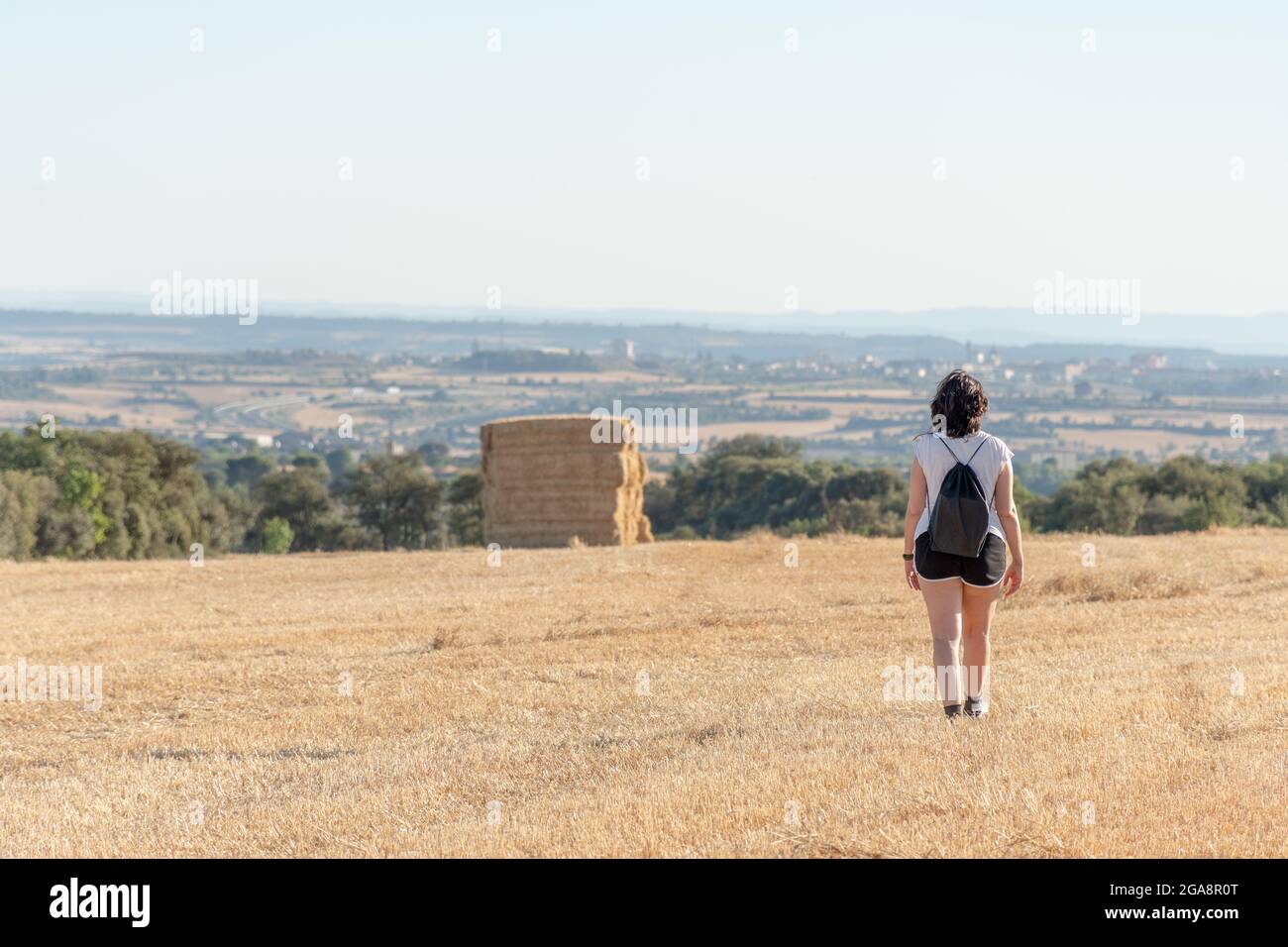 Eine Brünette mittleren Alters, die Shorts und ein weißes T-Shirt trägt, mit einem schwarzen Rucksack auf dem Rücken, der durch ein geerntetes Feld läuft. Strohhaufen Stockfoto