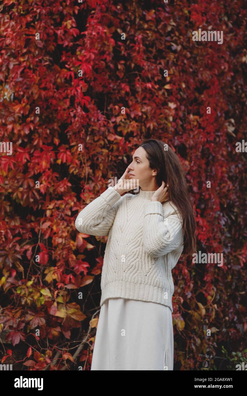 Langhaarige Frau in weißem Pullover, die im Herbst neben der roten Weintraubenwand steht. Stockfoto