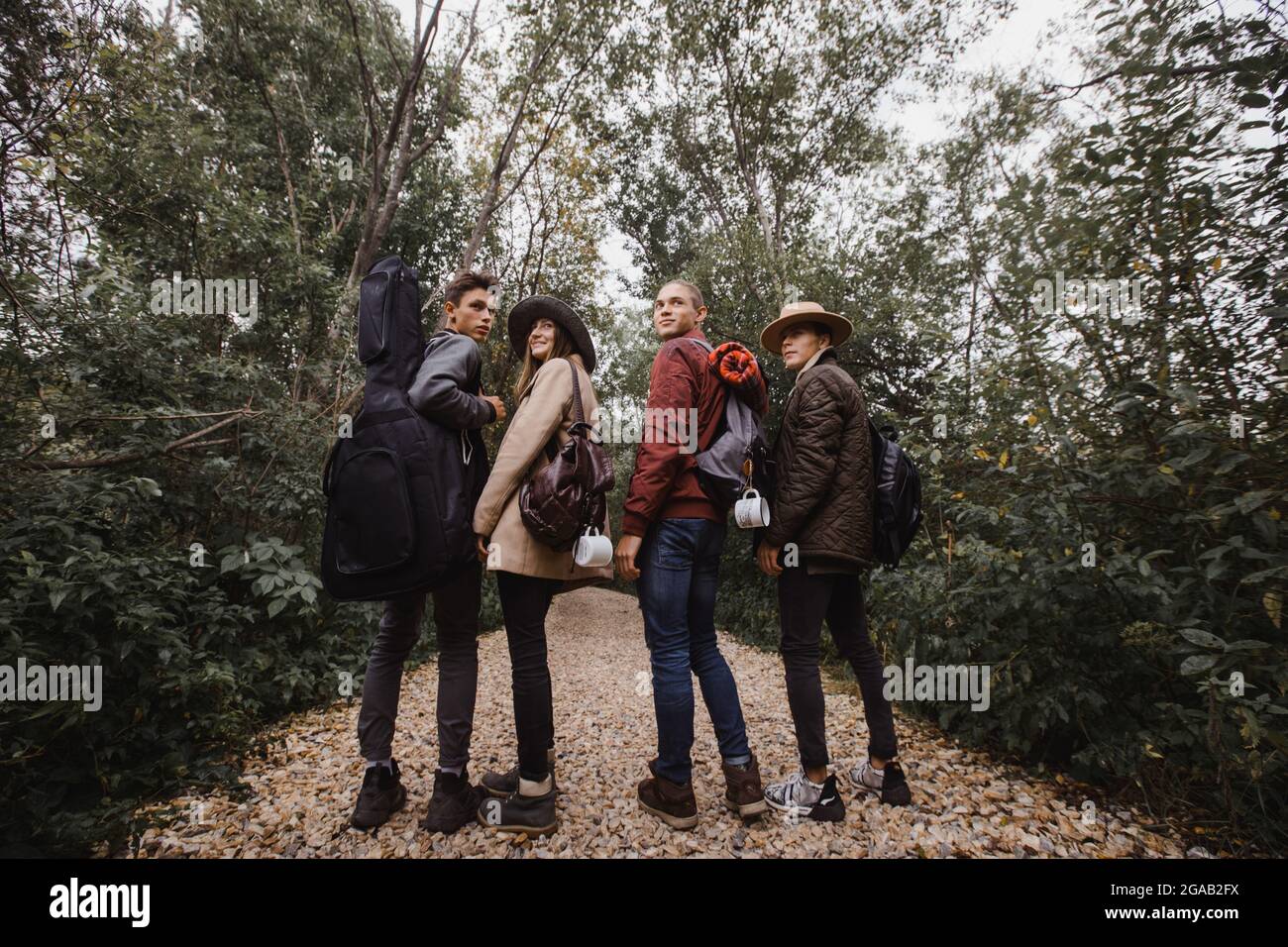 Vier junge Menschen in warmer Kleidung mit Gitarre und Rucksäcken stehen in einem Hain mit grünen Bäumen und blicken zurück Stockfoto Vier junge Menschen in warmer Kleidung mit Gitarre und Rucksäcken stehen in einem Hain mit grünen Bäumen und blicken zurück Stockfoto