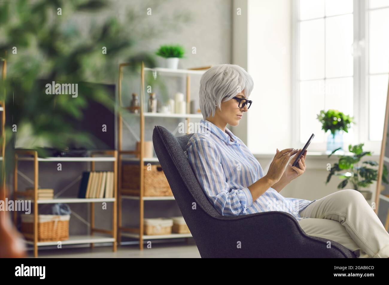 Seriöse ältere Frau, die im Wohnzimmer im Sessel sitzt und ihr Mobiltelefon benutzt Stockfoto
