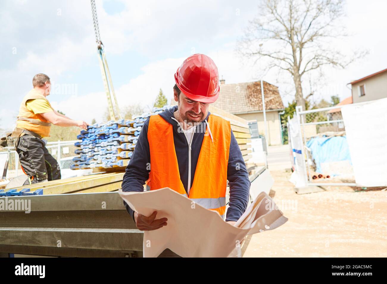 Architekt oder Vorarbeiter sieht sich eine Blaupause an, wobei die Lieferung von Baumaterial im Hintergrund steht Stockfoto