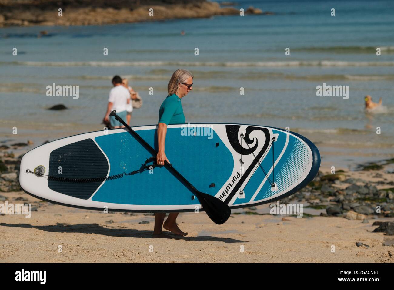 Frau mittleren Alters mit Paddleboard, die am Porthmeor Beach, St. Ives, Cornwall, Großbritannien, zum Meer läuft. Stockfoto