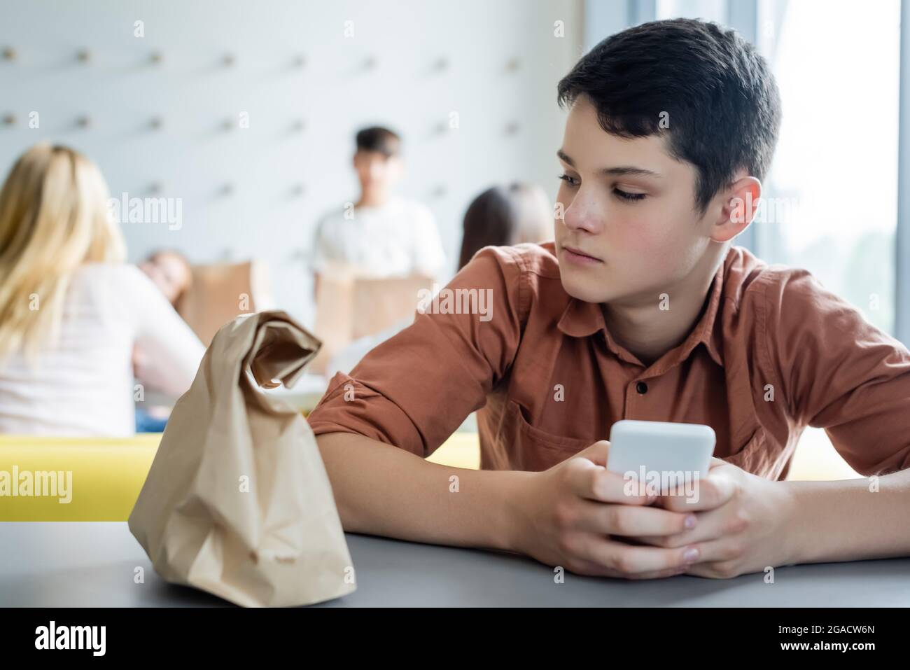 Trauriger Junge mit Mobiltelefon, das allein in der Schule in der Nähe einer Papiertüte sitzt, und verschwommenen Klassenkameraden Stockfoto