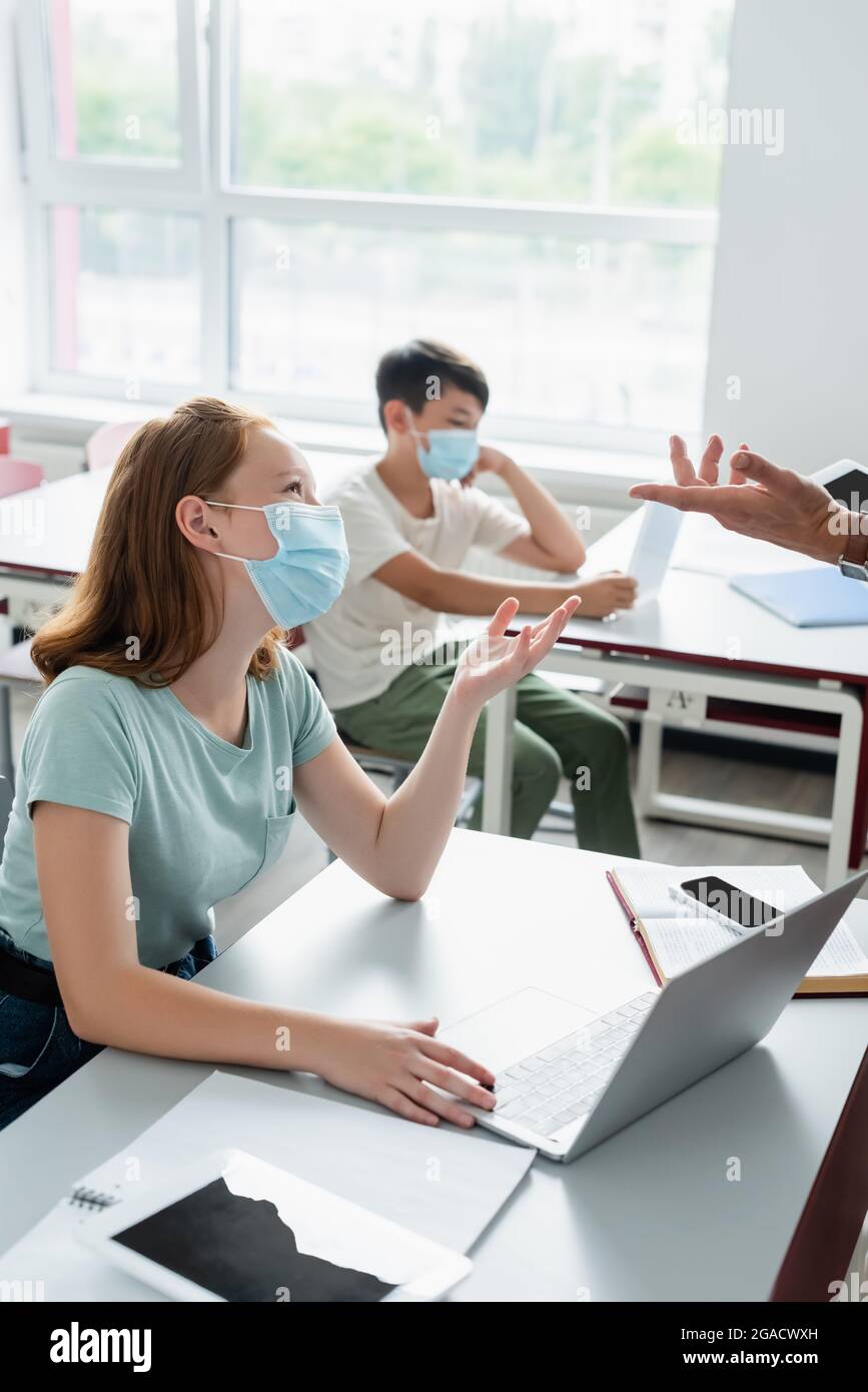 Schulmädchen in medizinischer Maske im Gespräch mit Lehrer mit digitalen Tablet während des Unterrichts Stockfoto
