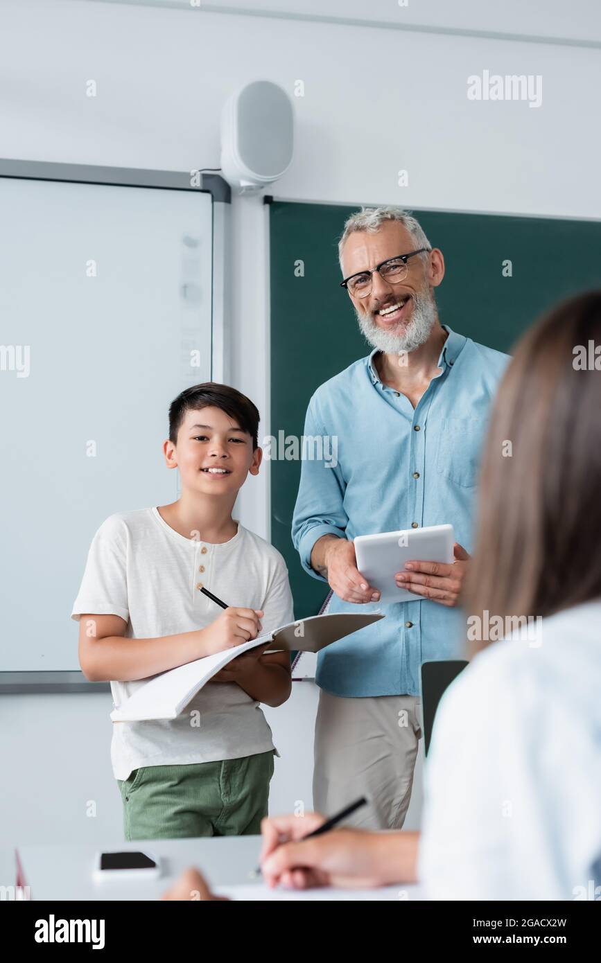 Lächelnder Lehrer mit digitalem Tablet, der die Kamera in der Nähe eines asiatischen Schülers mit Notebook anschaut Stockfoto