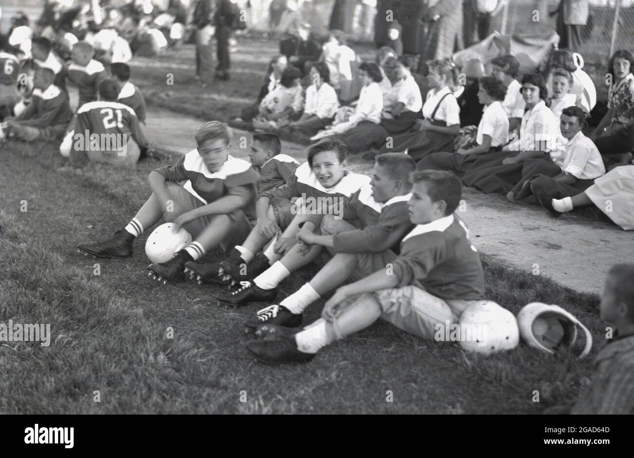 50er Jahre, historisch, High School, american Football, Schüler in ihrem Fußballtrikot, mit ihren Helmen sitzen zusammen draußen auf dem Boden, mit Schülerinnen sitzen hinter in ihren Uniformen dort zur Unterstützung, USA. Stockfoto