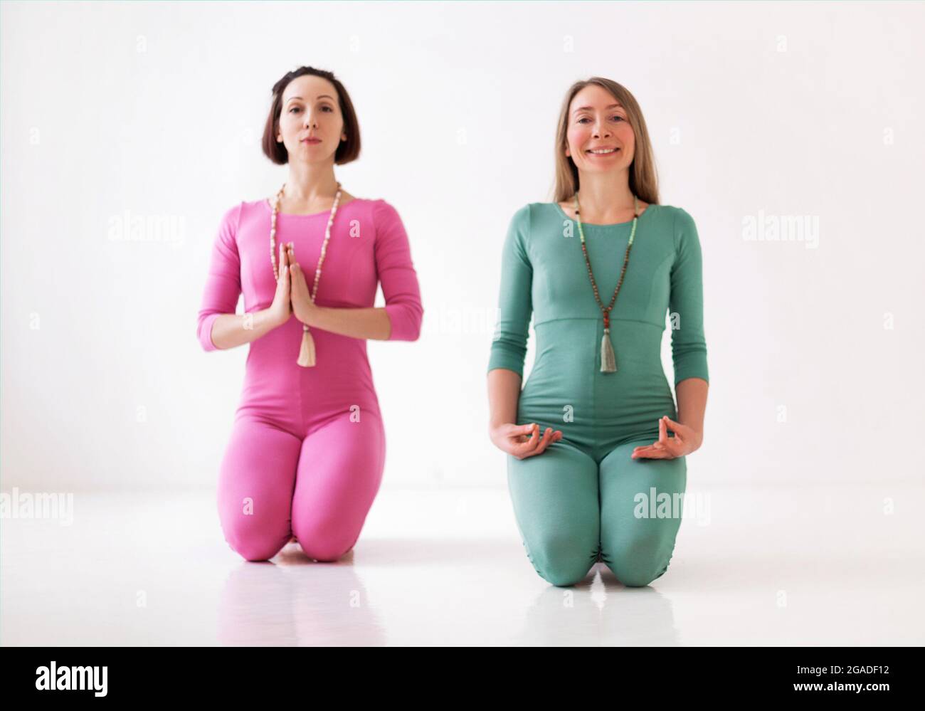 Zwei junge, glückliche, gesunde Frauen in langen Sportanzügen, die zusammen Yoga-Asanas während eines ziemlich meditativen Trainings oder einer Gruppe Yoga-Klasse durchführen, isoliert Stockfoto
