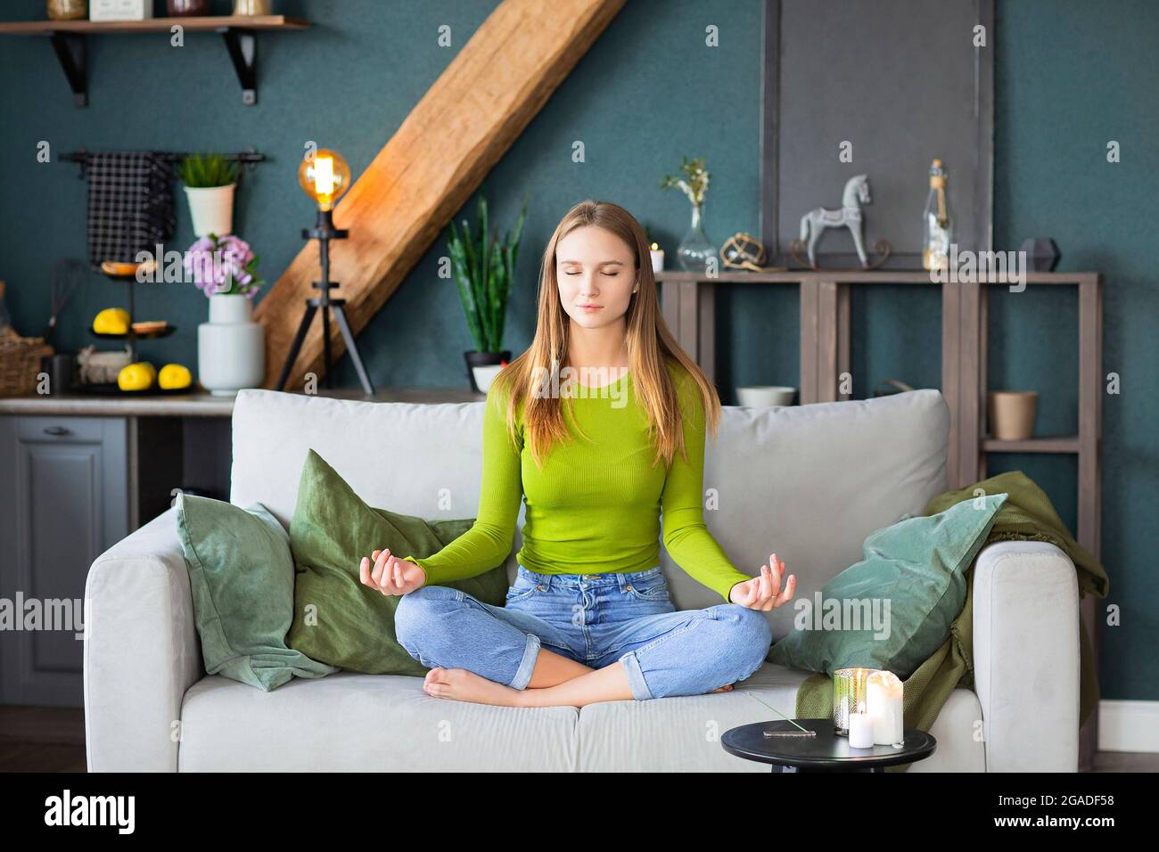Entspanntes, friedliches, tausendjähriges blondes Mädchen mit geschlossenen Augen, das in Lotuspose auf der Couch im Wohnzimmer zu Hause sitzt und eine Pause zur Meditation des Yoga macht Stockfoto