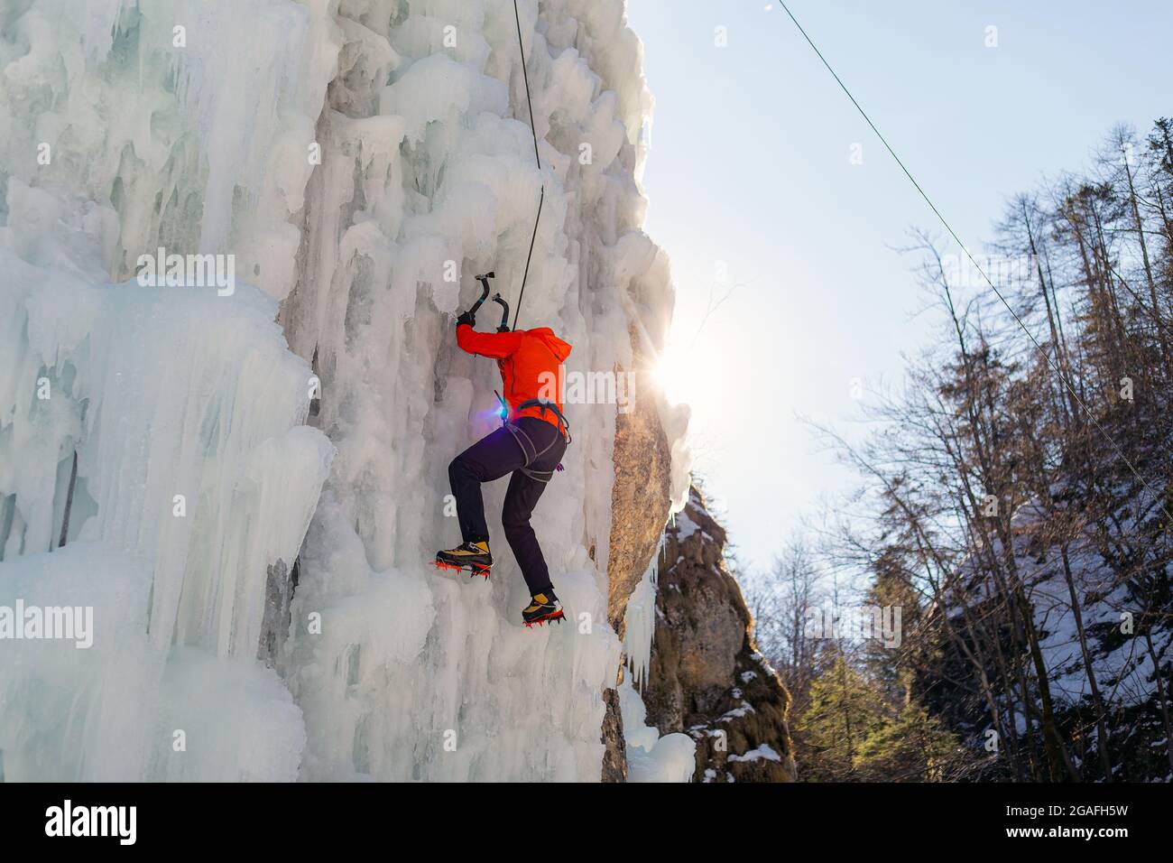 Luftaufnahme eines gefrorenen Eiswasserfalls, und der männliche Kletterer schreitet den Hang hinauf, während ein Seil, das an seinem Geschirr befestigt ist, ihn daran hindert, zu fallen Stockfoto