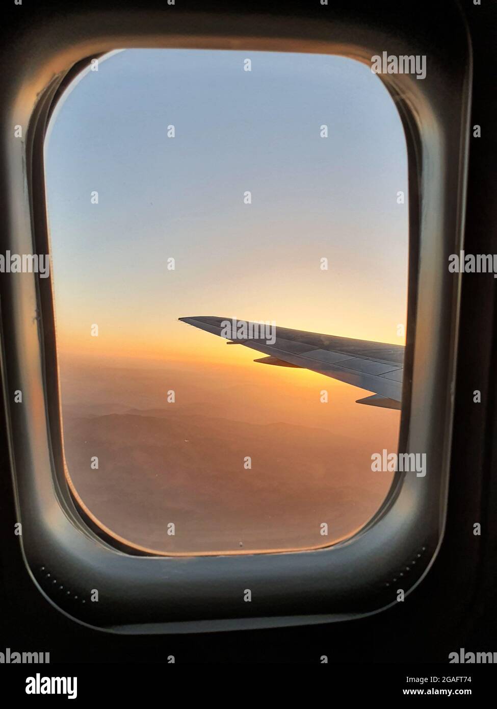 Blick auf den Himmel und den Flügel mit dem Iluminator des Flugzeugs. Schöner Sonnenuntergang, Berge und Reisetmosphäre. Stockfoto