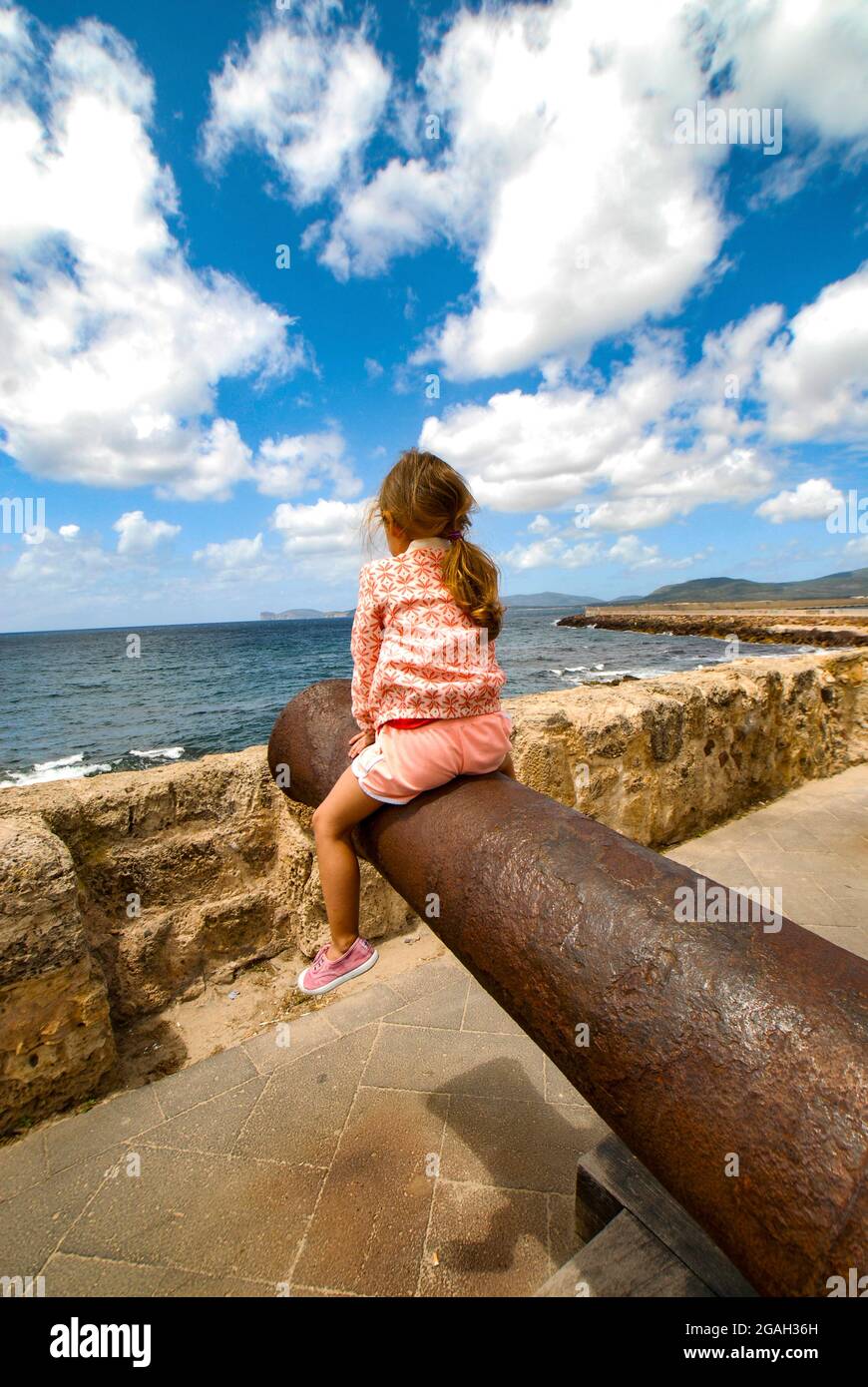 Alghero, ein kleines Mädchen, das auf einem alten Canno am Marco Polo Defensive Shield sitzt, kopiert ein mittelalterliches Katapult auf der Promenade, die auf die Uferpromenade der Insel Sard blickt Stockfoto