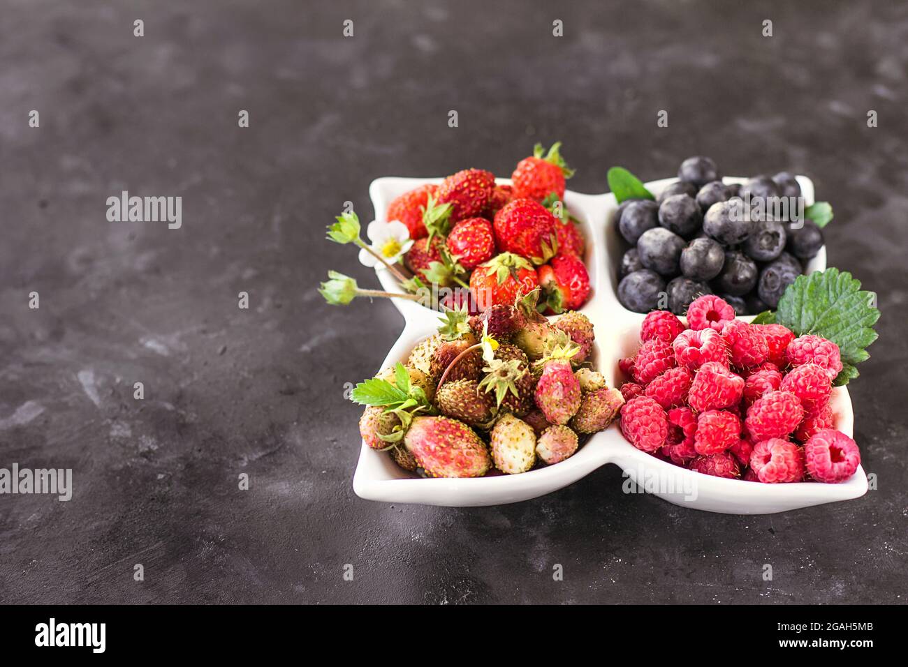 Eine Mischung aus frischen Beeren auf einem weißen Teller auf einem dunkelgrauen Hintergrund, natürliche Rohkost Stockfoto