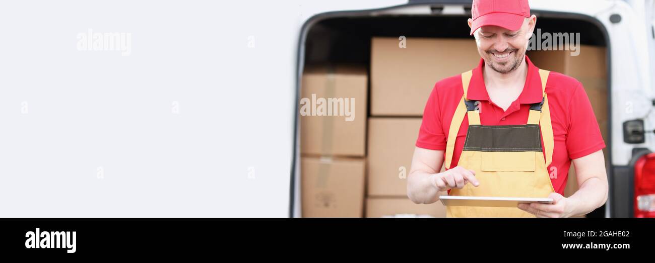 Männlicher Kurier in Uniform steht neben dem Auto mit Kisten und Hält das Tablet Stockfoto