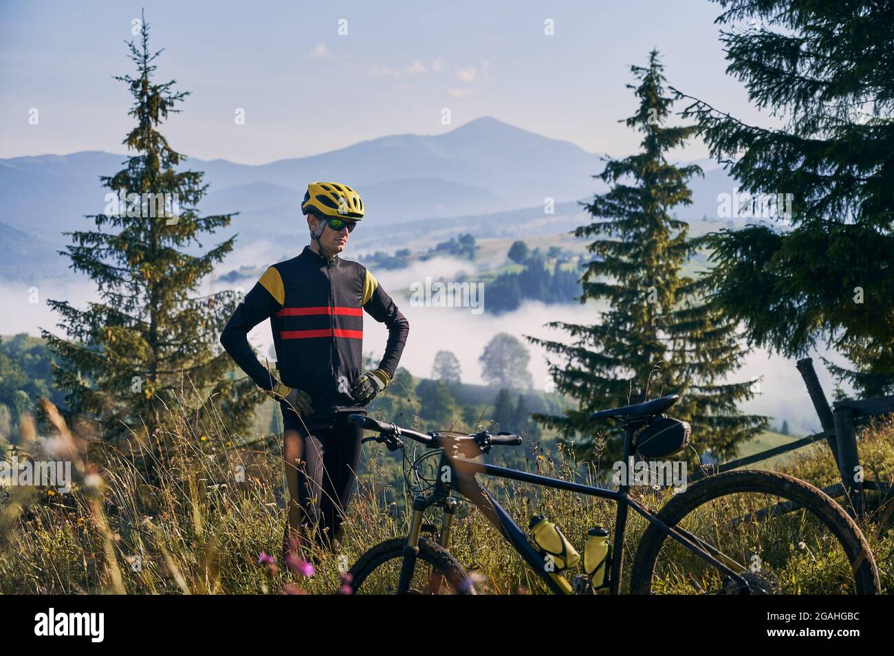 Radfahrer in Uniform und Helm, stehend auf einem grasbewachsenen Hügel. Schöne Berglandschaft im Hintergrund. Am frühen Morgen in den neblig sonnigen Bergen. Fahrrad auf verschwommenem Vordergrund. Stockfoto