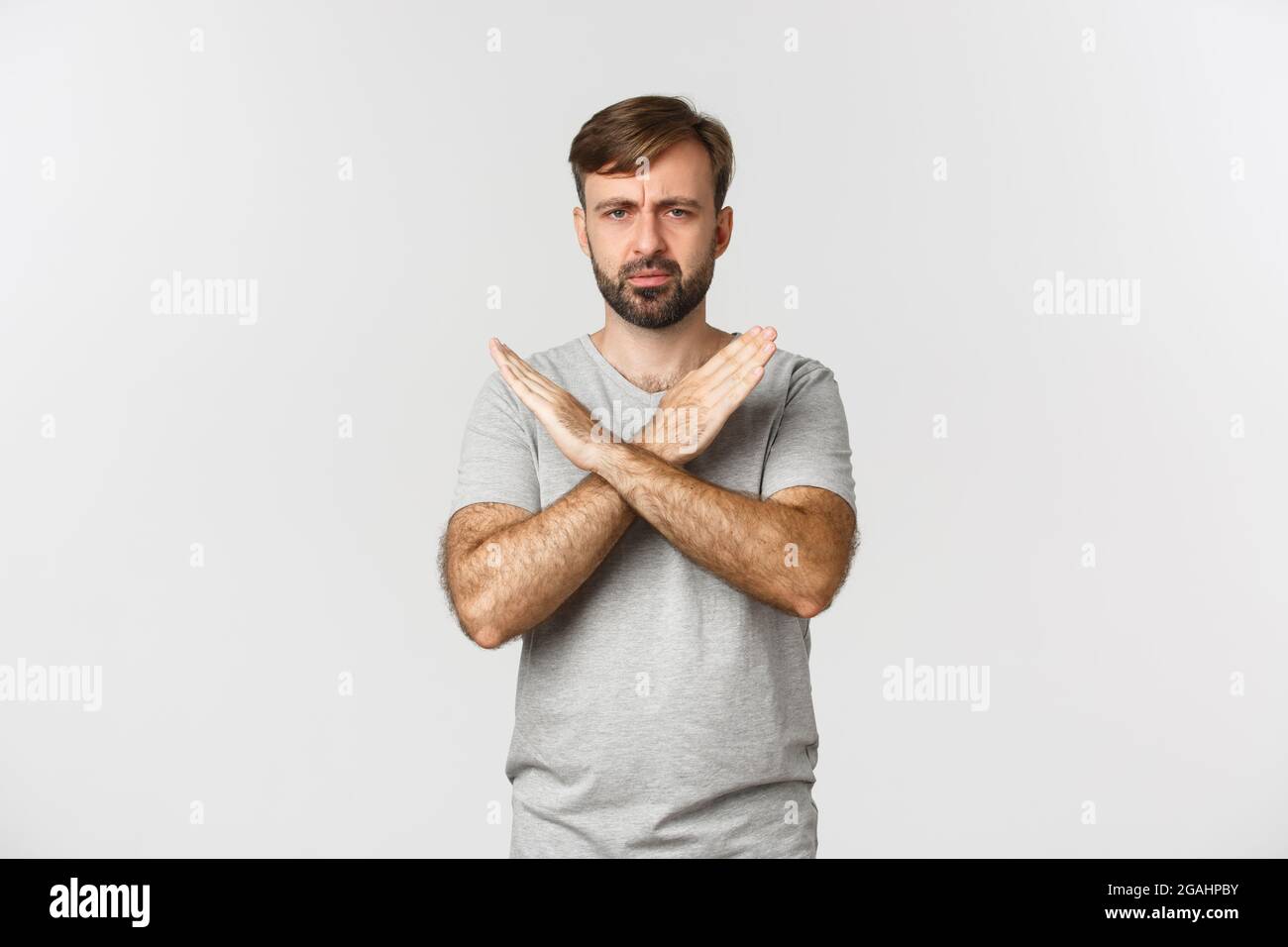 Enttäuschter Mann mit Bart, zeigt Kreuzgesten und Stirnrunzeln, sagt zu stoppen, verbieten etwas Schlechtes, stehen über weißem Hintergrund Stockfoto