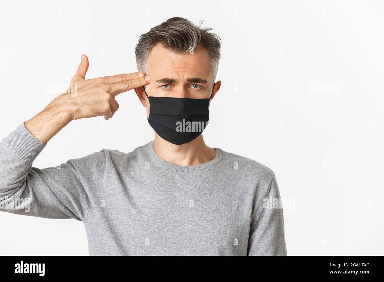 Konzept von covid-19, sozialer Distanzierung und Quarantäne. Nahaufnahme eines traurigen Mannes mittleren Alters in schwarzer medizinischer Maske, mit der Fingerpistole auf den Kopf zeigend und schauend Stockfoto