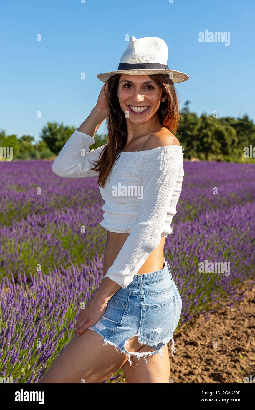 Halbkörperporträt einer lächelnden jungen Frau in einem blühenden Lavendelfeld. Lässiger Look mit kurzer Jeans-Hose, weißem Oberteil und Mütze. Sommerkonzept. Stockfoto