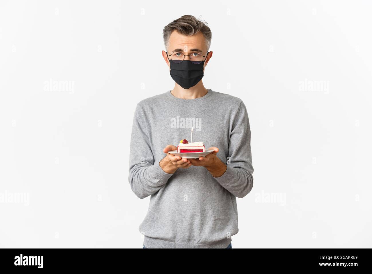 Konzept von Coronavirus, Quarantäne und Urlaub. Bild von traurigen Mann mittleren Alters in schwarzer medizinischer Maske und Brille, hält Geburtstagskuchen mit brennenden Kerze Stockfoto