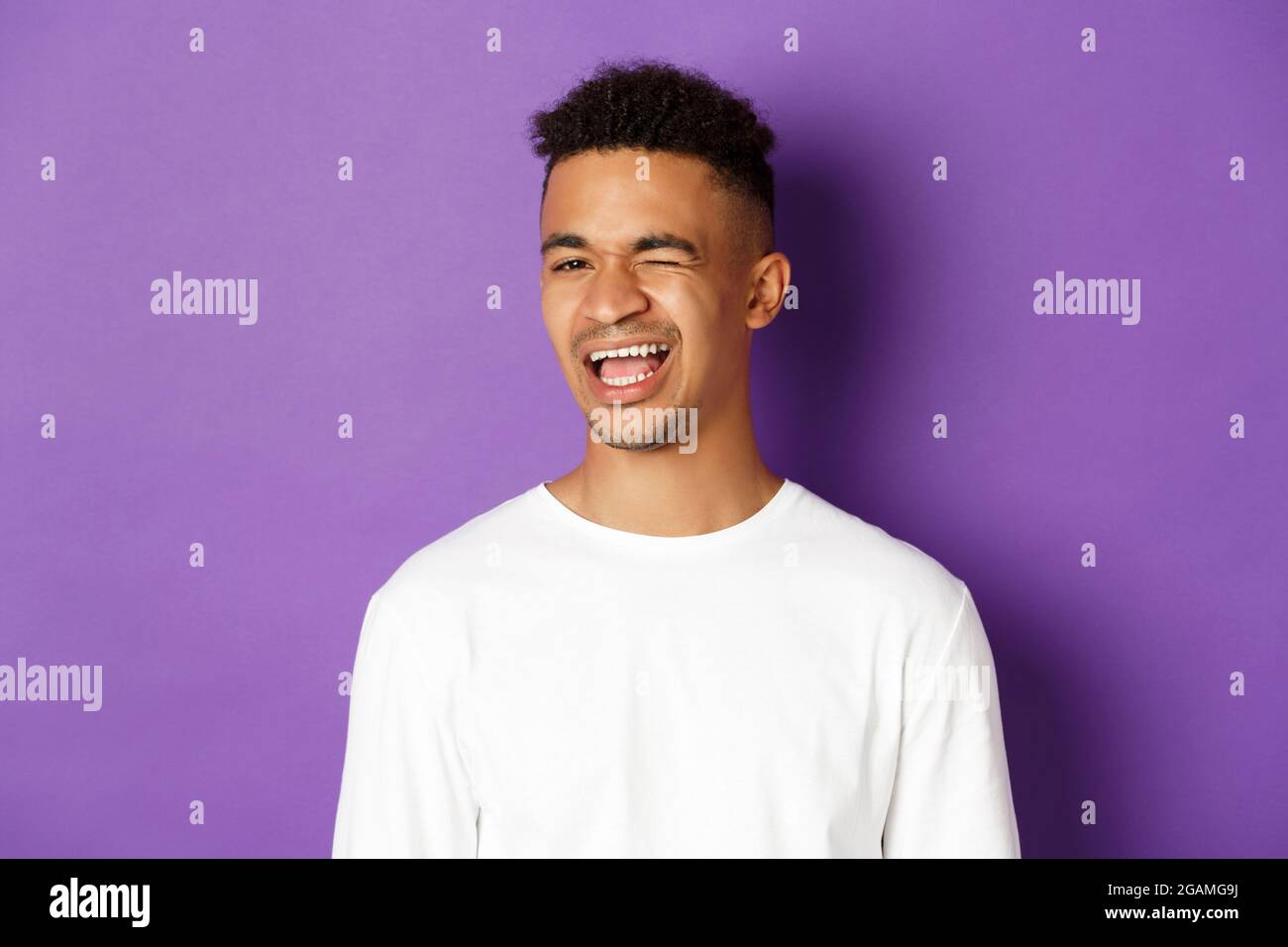Nahaufnahme eines hübschen jungen afroamerikanischen Mannes, der blinzelnd, lächelnd und fröhlich aussagt und auf purpurem Hintergrund steht Stockfoto