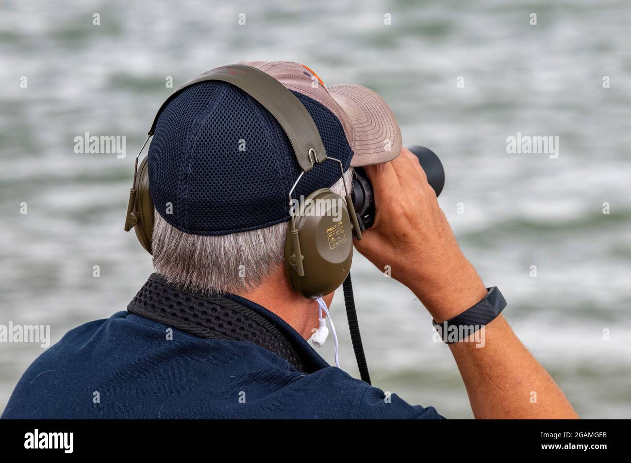 Älterer Mann mit Fernglas, Rentner, der durch ein Fernglas schaute, Mann mit Fernglas, Mann mit Ohrenschützern, Zuschauer bei der cowes Week, Spionage. Stockfoto