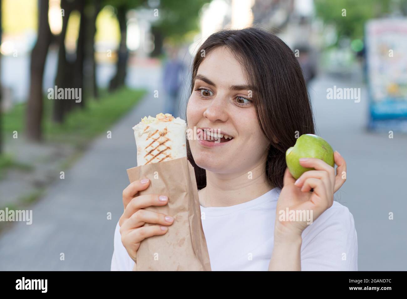 Junge schöne Brünette Frau auf einer Diät wählt Nahrung - was einen Apfel oder Fast Food zu essen. Beißt das Shawarma und wählt die Völlerei Stockfoto