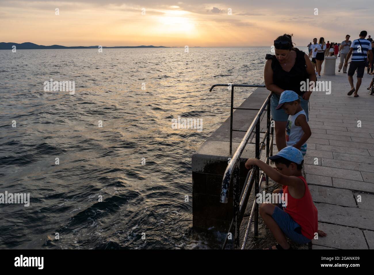 ZADAR, KROATIEN - 21. Jul 2021: Die Kinder, die bei Sonnenuntergang in Zadar, Kroatien, an einer Metallbarriere in der Nähe des Wassers stehen Stockfoto