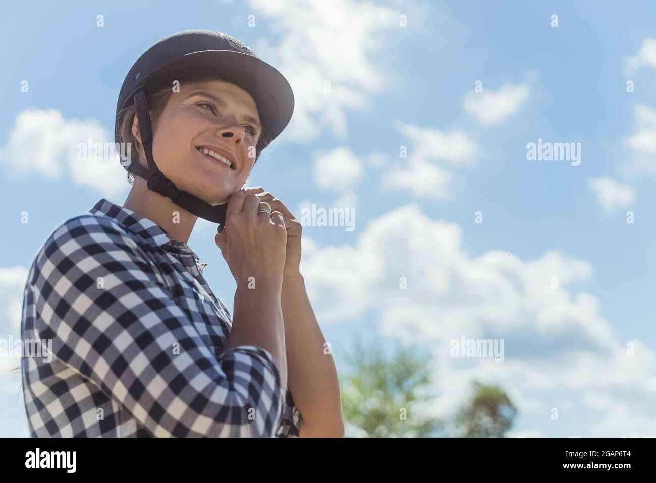 Weibliche Jockey, die einen Helm aufsetzt und sich für den Reitwettbewerb vorbereitet. Eine wettbewerbsfähige Frau, die alle auf den Wettbewerb eingestellt ist. Konzept des Reitwettbewerbs. Wolkiger Himmel im Hintergrund. Stockfoto