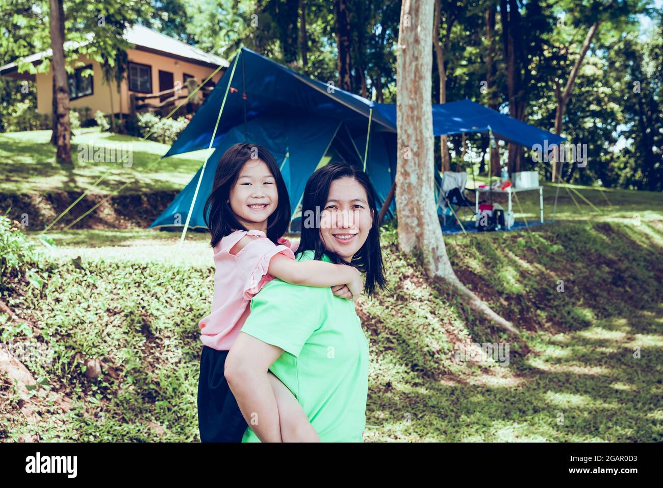 Süße asiatische Tochter auf einem Schweinchen-Rückritt mit ihrer Mutter im Nationalpark mit Camping-Zelt auf dem Hintergrund. Stockfoto