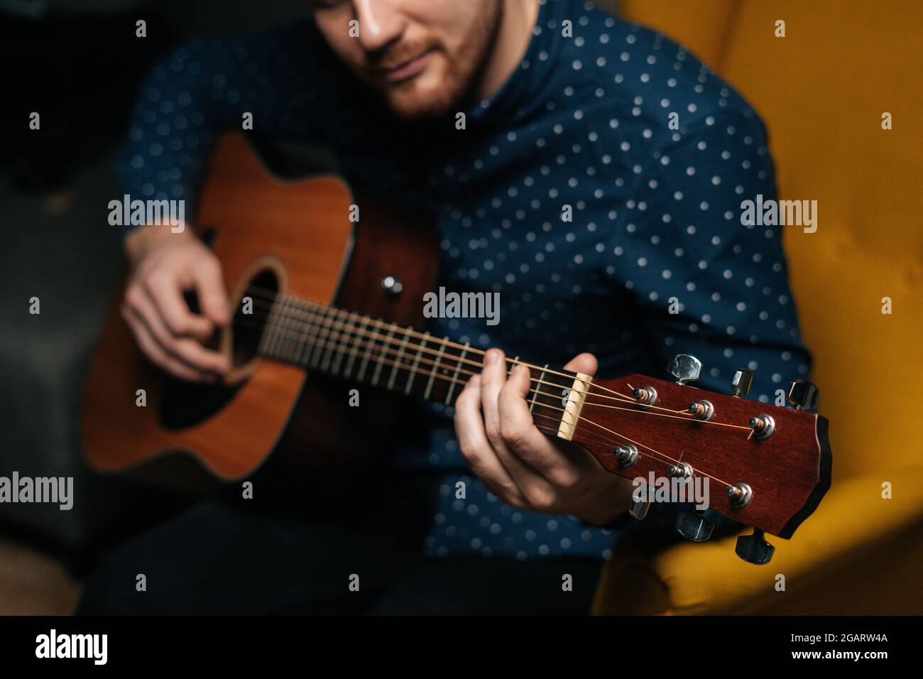 Nahaufnahme des Gitarristen, Sänger, der akustische Gitarre spielt, sitzt auf einem Sessel im dunklen Wohnzimmer, selektiver Fokus. Stockfoto