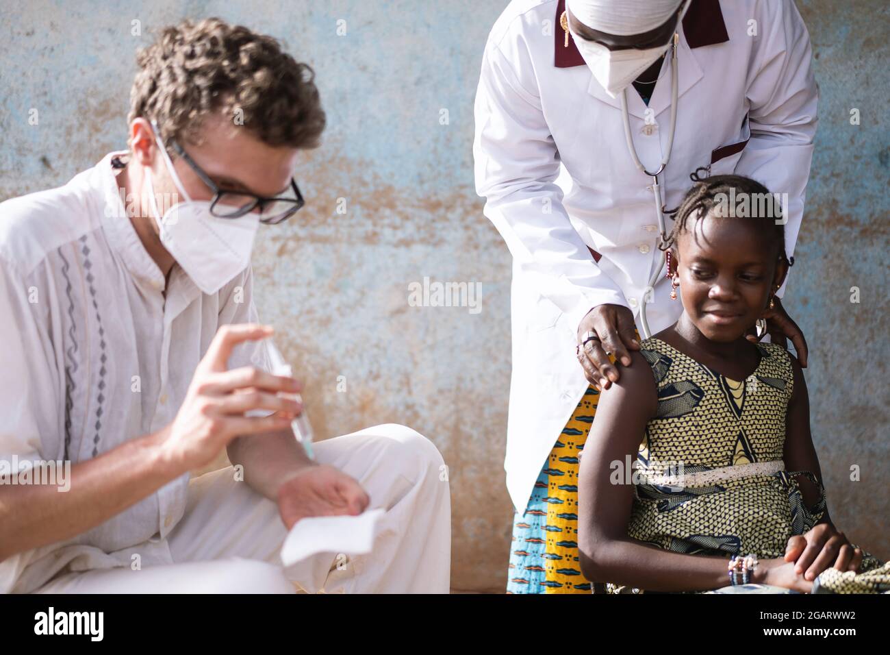 In diesem Bild bereitet ein kaukasischer Arzt eine Spritze vor, während ein selbstbewusstes kleines afrikanisches Mädchen, das auf ihre Injektion wartet, von einem Sanftmütigen beruhigt wird Stockfoto