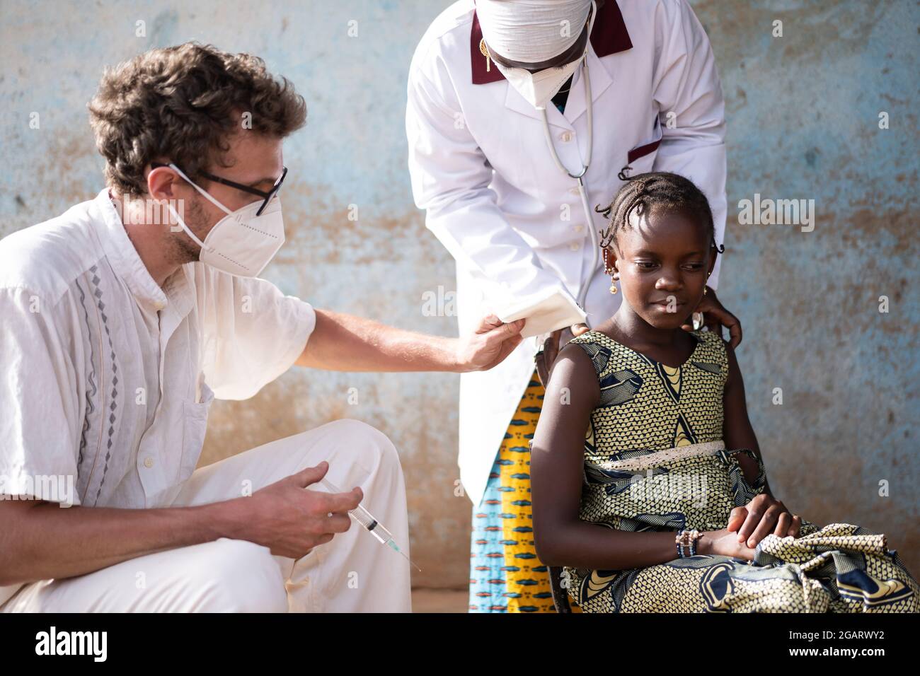 In diesem Bild desinfiziert ein kaukasischer Arzt mit Gesichtsmaske die Injektionsstelle am Arm eines mutigen kleinen afrikanischen Mädchens, das von einem bla ermutigt wird Stockfoto