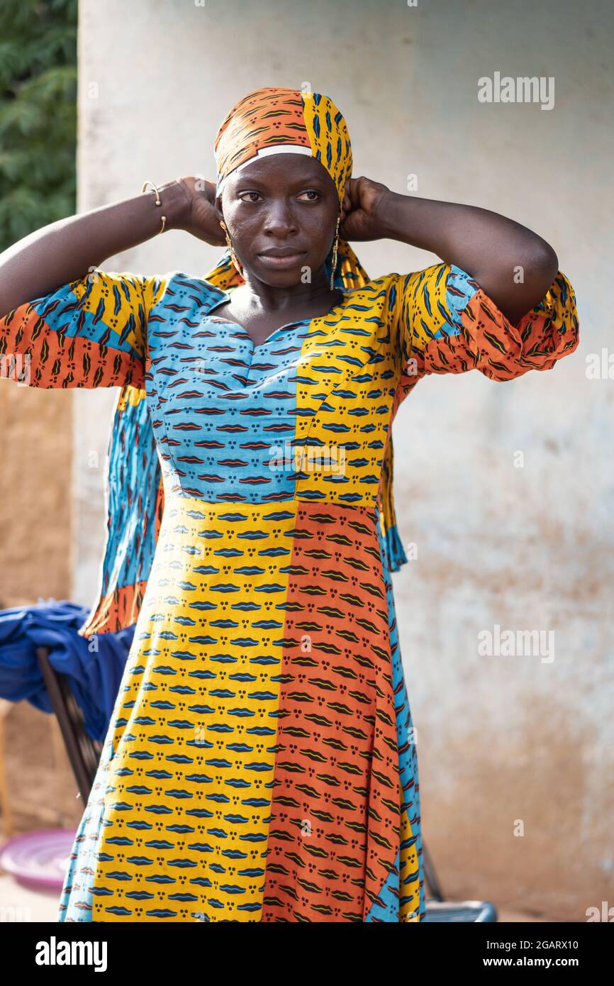 In diesem Bild versucht eine nachdenkliche junge schwarz-afrikanische Schönheit mit erhobenen Armen, ihre Kopfbedeckung anzupassen, die zu ihrem kolossal traditionellen Kleid passt Stockfoto
