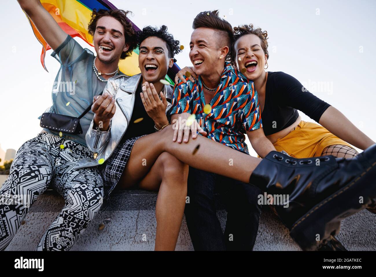 Junge Menschen feiern Stolz, während sie zusammen sitzen. Vier Mitglieder der LGBTQ+ Community lächeln fröhlich und heben die Pride-Flagge. Gruppe von Stockfoto