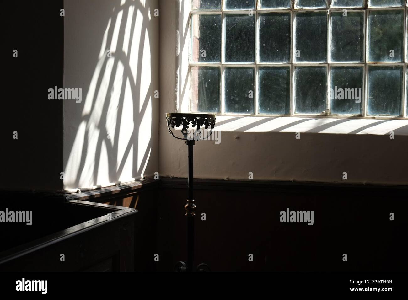 Das Licht der Fensterscheiben wirft unregelmäßige geometrische Schatten auf eine Kirchenwand über der Vertäfelung. Ein Messing Blume Schüssel Halter ist silhouetted Stockfoto