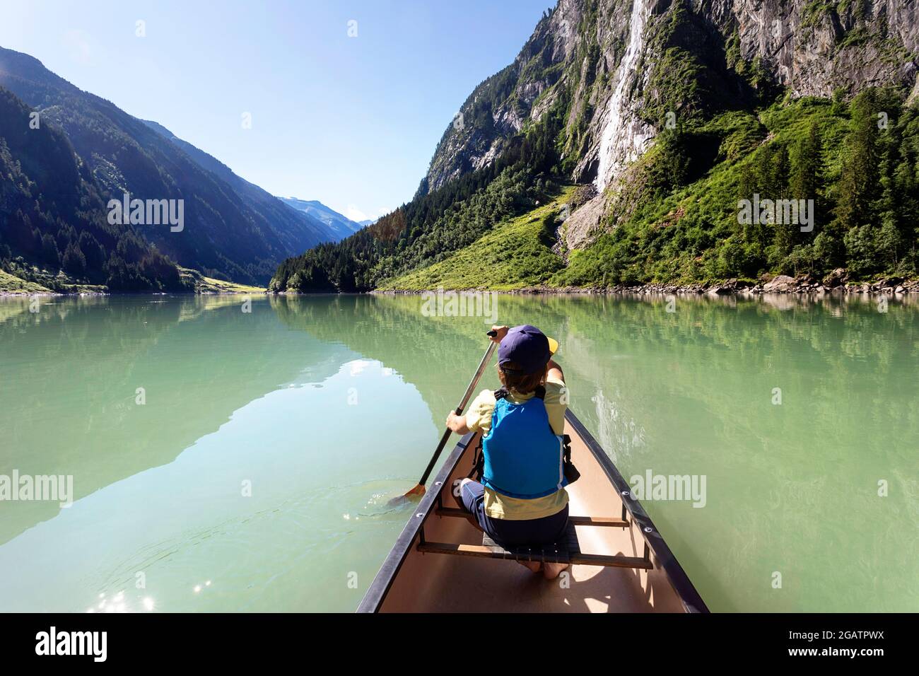 Junge sitzt im Kanu Reiten auf Bergsee in Sommer alpine Landschaft mit Wasserfall in Bacgrund. Stillup Stausee, Stillup Lake, Österreich, Tirol Stockfoto