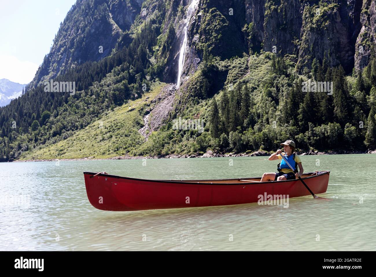 Junge sitzt im Kanu Reiten auf Bergsee in Sommer alpine Landschaft mit Wasserfall in Bacgrund. Stillup Stausee, Stillup Lake, Österreich, Tirol Stockfoto