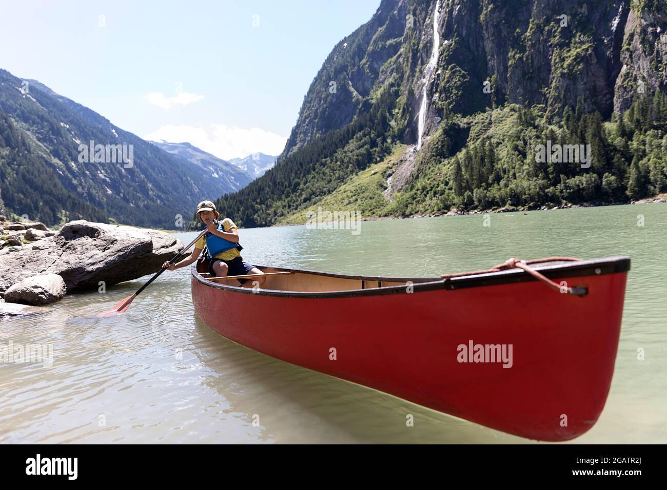 Junge sitzt im Kanu Reiten auf Bergsee in Sommer alpine Landschaft mit Wasserfall in Bacgrund. Stillup Stausee, Stillup Lake, Österreich, Tirol Stockfoto