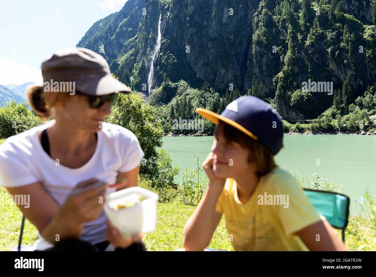 Mutter und Sohn beim Mittagessen an einem Bergsee in der sommerlichen Alpenlandschaft mit Wasserfall im Hintergrund. Stillup Stausee, Stillup Lake, Österreich, Tirol Stockfoto