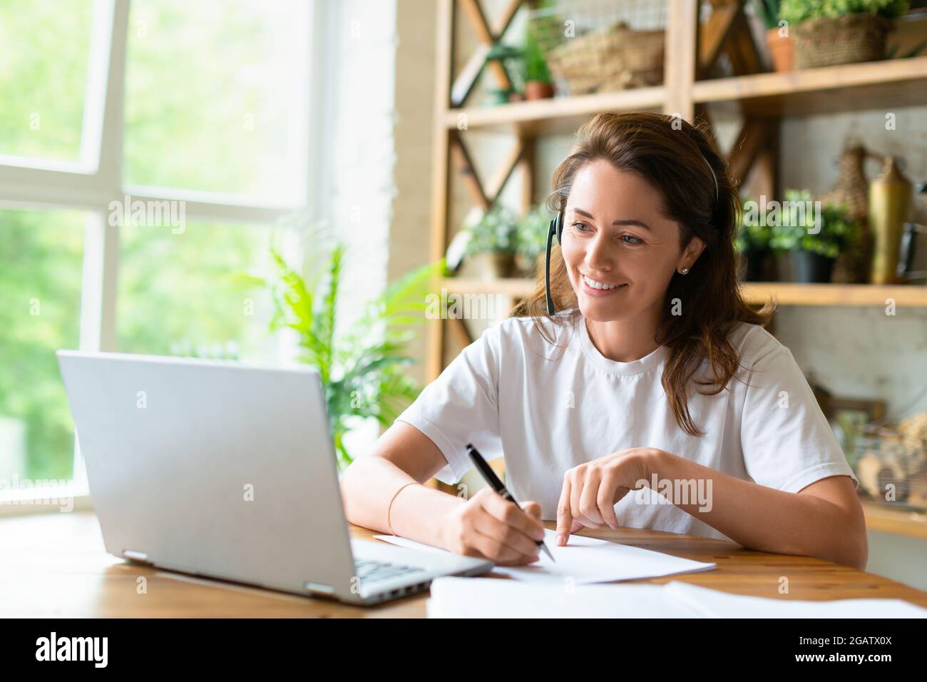 Eine lächelnde Frau arbeitet zu Hause vor einem Laptop-Monitor mit Papieren. Während eines Online-Videoanrufs prüft sie die Papiere. Modernes Technologiekonzept. Stockfoto