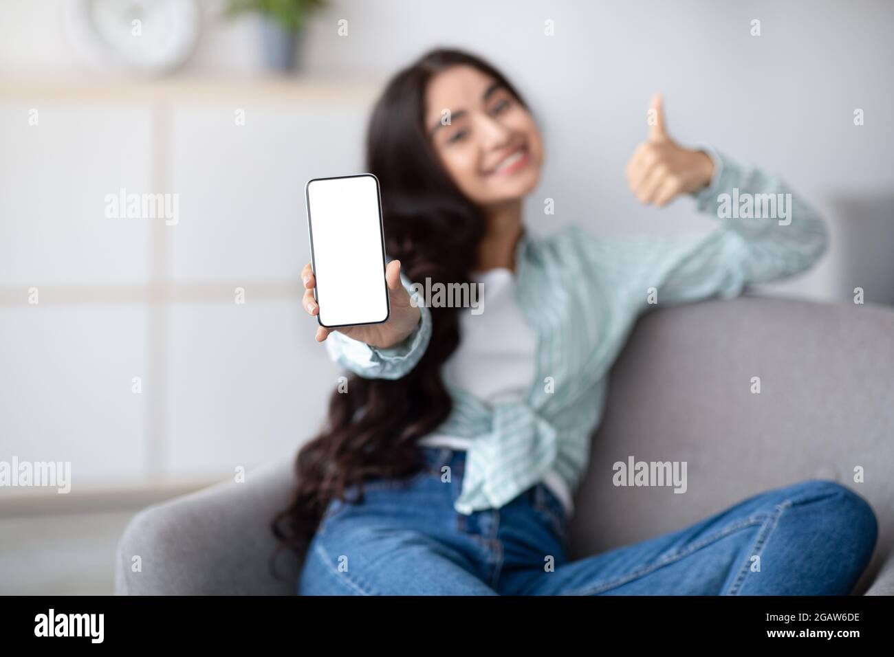 Sieh dir diese tolle mobile App an. Hübsche indische Dame präsentiert Smartphone mit weißem leeren Bildschirm und zeigt Daumen nach oben zu Hause, Mockup für Website o Stockfoto
