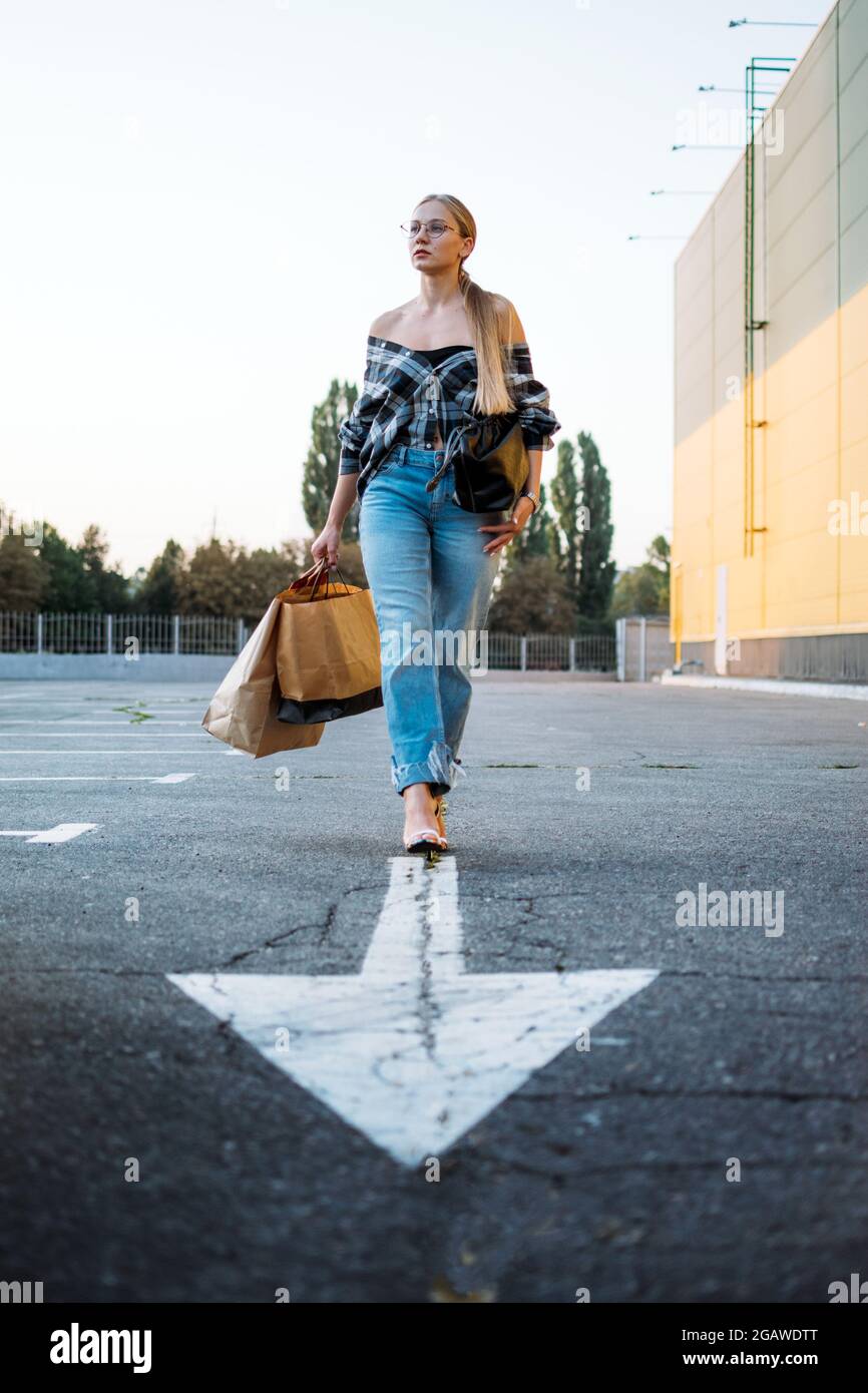 Shopping, Verkauf, Happy Young stilvolle blonde Frau mit Einkaufstaschen gehen zum Auto auf dem Parkplatz auf weißem Pfeil nach dem Einkaufen. Frau mit Einkaufstaschen Stockfoto