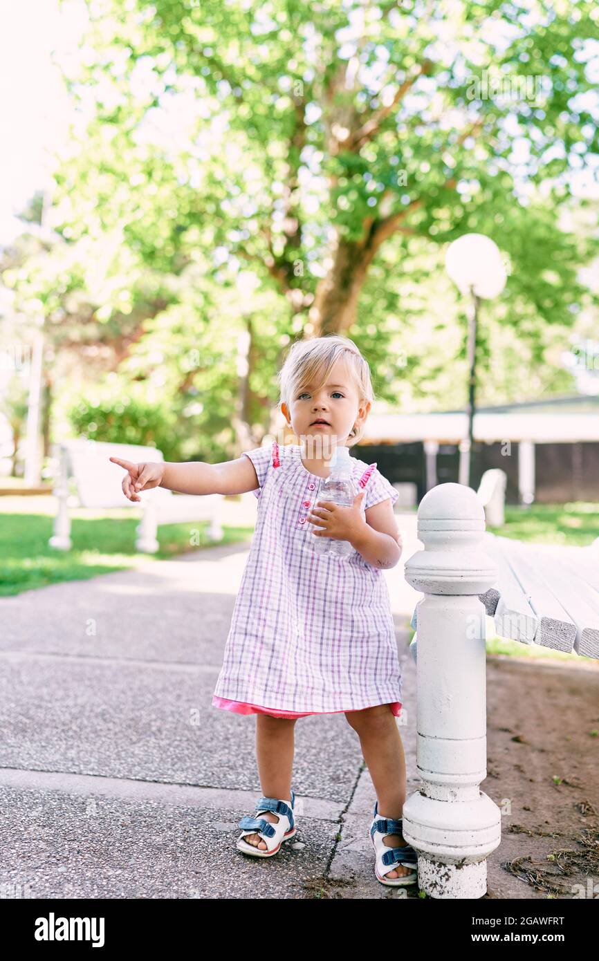 Das kleine Mädchen mit einer Flasche Wasser in der Hand steht an einer  Parkbank und zeigt ihren Finger zur Seite Stockfotografie - Alamy