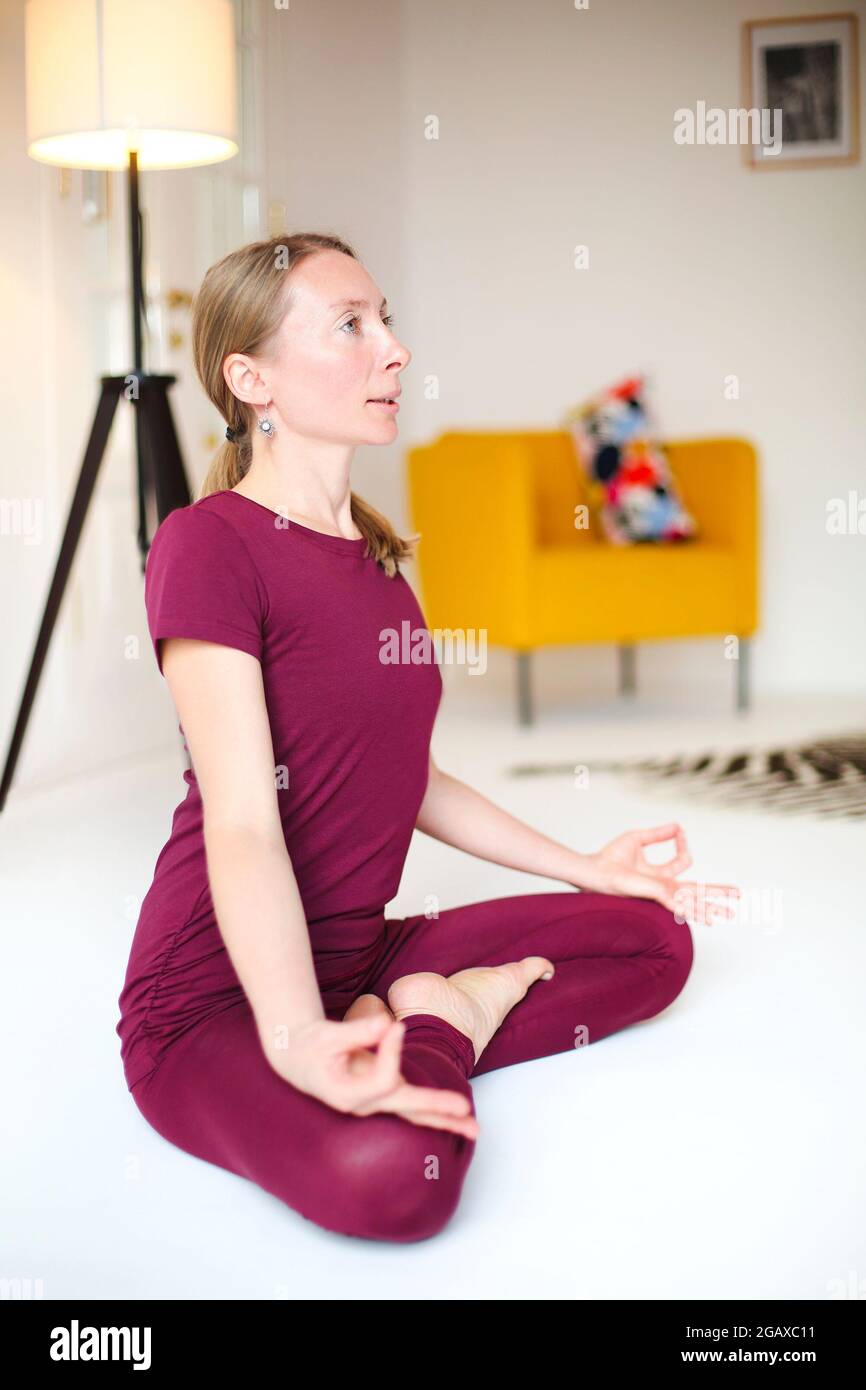 Blonde Frau in Sportkleidung sitzt mit gekreuzten Beinen und Mudra-Händen auf der Matte und macht Yoga, während sie Achtsamkeit übt Stockfoto