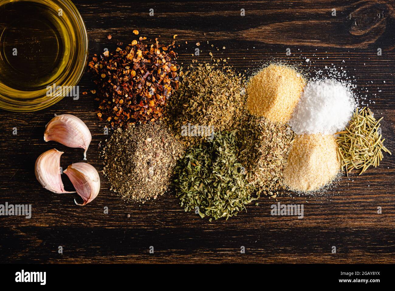 Zutaten für italienisches Brot auf einem Holztisch: Getrocknete Gewürze, Knoblauchzehen und natives Olivenöl extra auf dunklem Holzboden Stockfoto