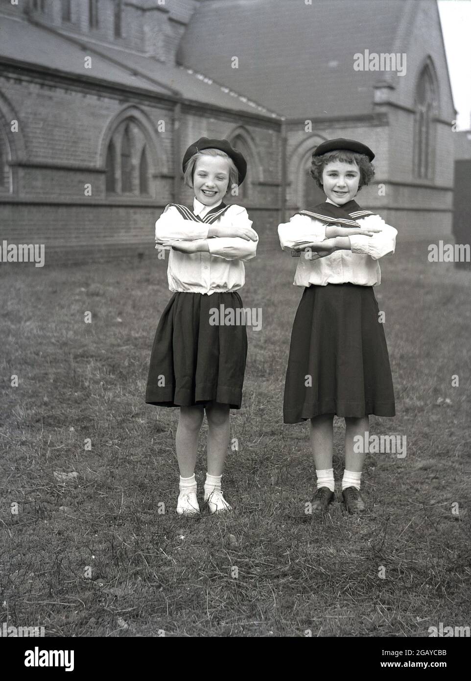 1956, historisch, draußen auf dem Gelände einer Kirche, zwei Mädchen, die in ihren Kostümen und Barets für das besondere 1. Mai-Karneval stehen, mit ihren Händen auf ihren Armen gefaltet, Leeds, England, Großbritannien. Stockfoto