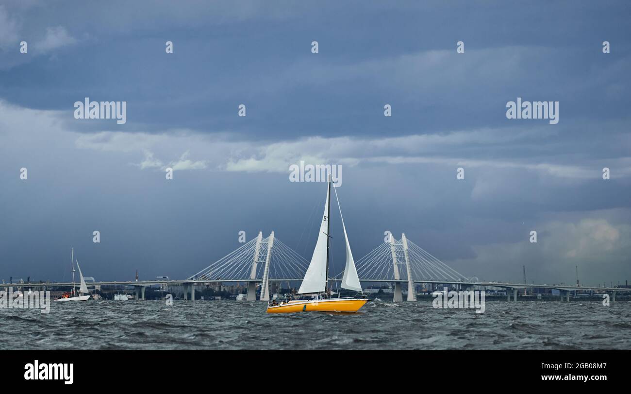 Das gelbe Segelboot am Horizont im Meer bei Sonnenuntergang, Kabelbrücke im Hintergrund, der Sturmhimmel in verschiedenen Farben, große Wellen, Segelregatta, bewölkt Stockfoto