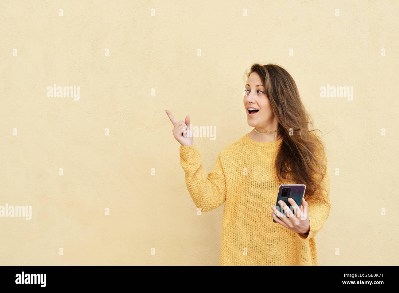 Frau mit einer aufgeschreckte Geste und einem Smartphone, das auf einen gelben Hintergrund zeigt, um Platz zu kopieren Stockfoto