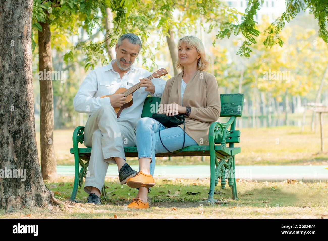 Happy Elder Liebe Paar in Romantik sitzen und spielen Ukulele Musikinstrument auf Stuhl im Park Stockfoto